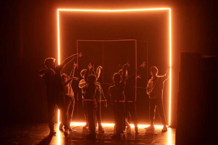 Group of people raising their hands inside a glowing orange neon square on stage.