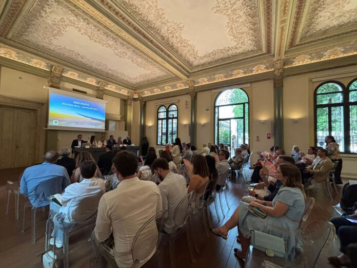 Panel of speakers presenting at a formal conference in a historic, frescoed room with an audience seated before them.