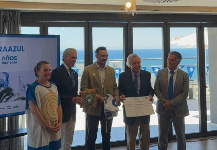 Five people stand indoors at an awards ceremony; a man holds a wooden plaque and another a certificate, with a banner and ocean view visible through the windows behind them.