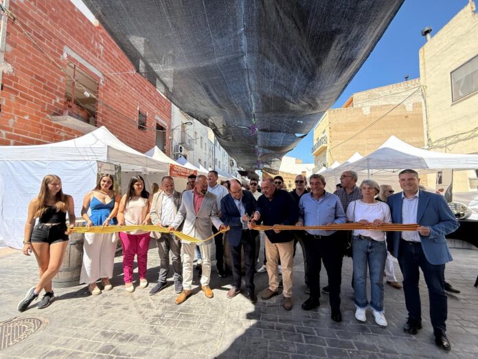 Group of people cutting an orange ribbon at an outdoor market or street opening, under a black shade canopy with white tents lining the street.