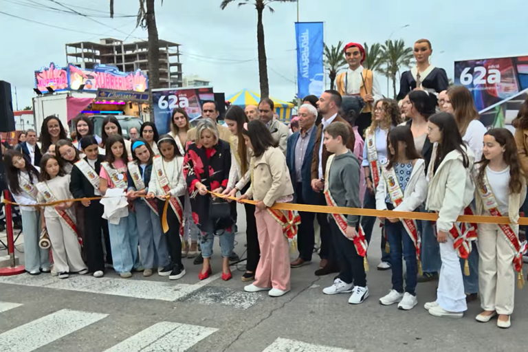 Group of children and adults at a ribbon-cutting ceremony on a city street, many wearing sashes and white outfits, cutting an orange ribbon.