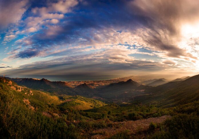 Panoramic view of rocky hills and green vegetation leading to a distant coastline and calm sea under a colorful sunset sky with scattered clouds.