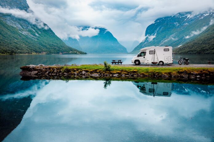 White camper van parked on a lakeside edge with a picnic table and bike nearby, surrounded by misty alpine mountains and a calm lake reflection.