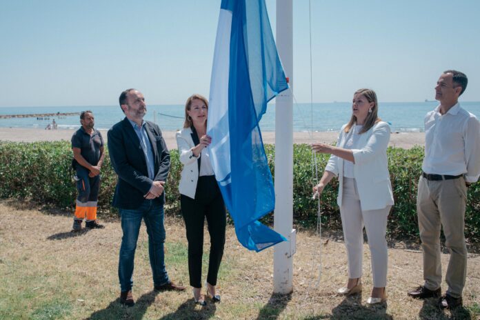 Five adults at the beach watch as a blue flag is raised on a white flagpole during a seaside ceremony.