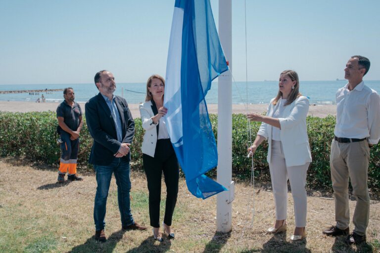 Five adults at the beach watch as a blue flag is raised on a white flagpole during a seaside ceremony.