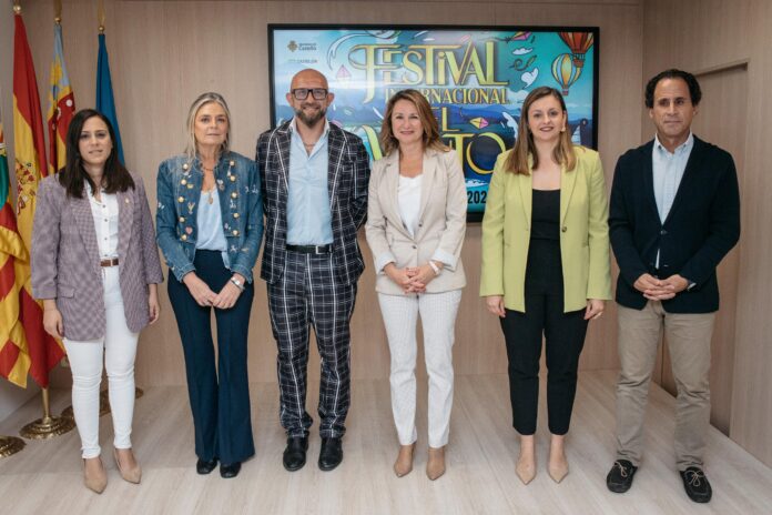 Six adults in smart-casual attire stand side by side in front of a colorful festival banner and flags, posing for a group photo in a formal setting.