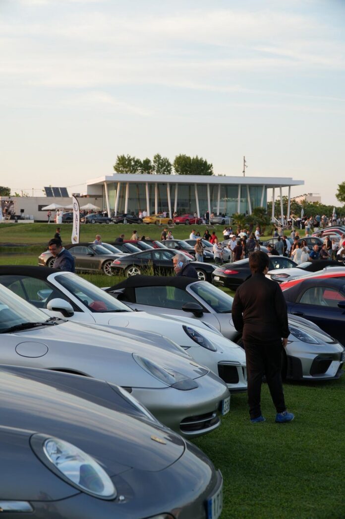 Line of silver sports cars parked on a grassy field with a crowd of people at a car show, modern glass building in the background.