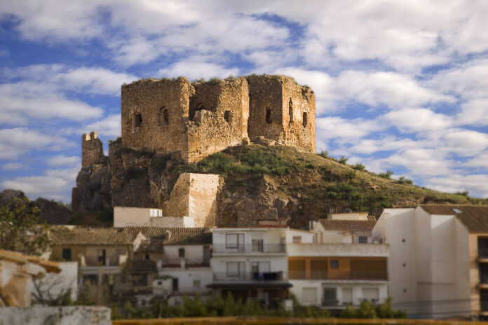 Ruined stone fortress atop a hill overlooking white houses against a blue sky with scattered clouds.