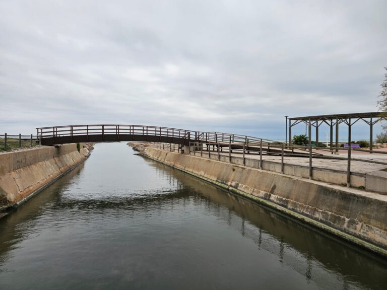 Wooden pedestrian bridge arches over a narrow canal with concrete banks and a cloudy sky; a wooden shelter/trellis is visible on the right.