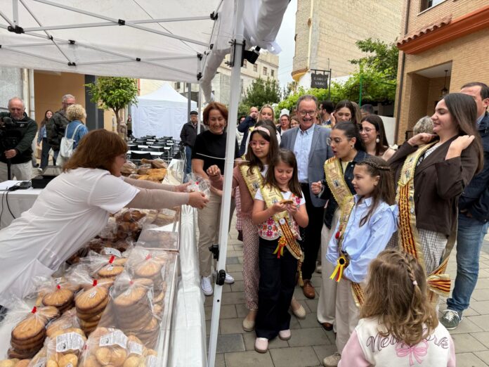 Woman hands out pastries from a bakery stall at an outdoor market while a group of children with sashes and adults watch.