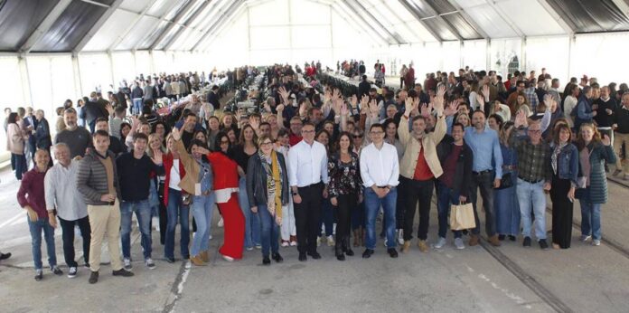 Large diverse group inside a bright, long tent waving and smiling at the camera, forming a front row.