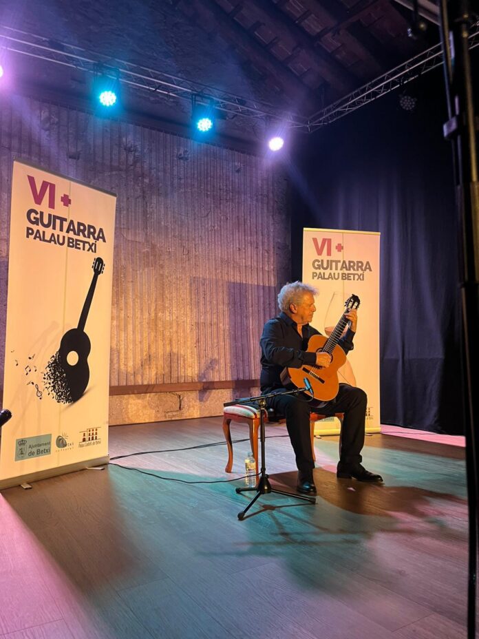 Male guitarist seated on stage playing a classical guitar, with VI+ Guitarra Palau Betxi banners beside him under colorful lights.