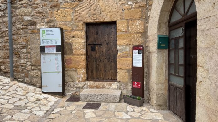 Stone building exterior with a closed wooden door, a tall metal map/info panel, and a small plant pot near an arched doorway.