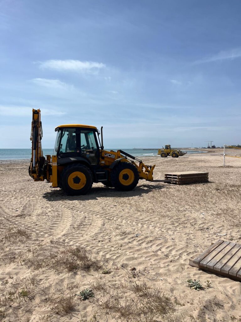 Yellow backhoe loader on a sandy beach with a stack of wooden pallets nearby, ocean in the background.