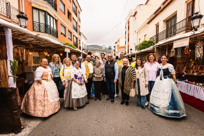Group of people posing in a street market lined with stalls and string lights, several women wearing traditional Valencian-style dresses and colorful outfits, hillside in the distance.