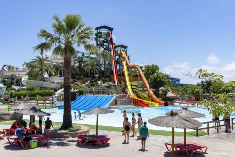 Sunny water park with tall multi-colored slides and twisting tubes, pool below, surrounded by palm trees and sunbathers.