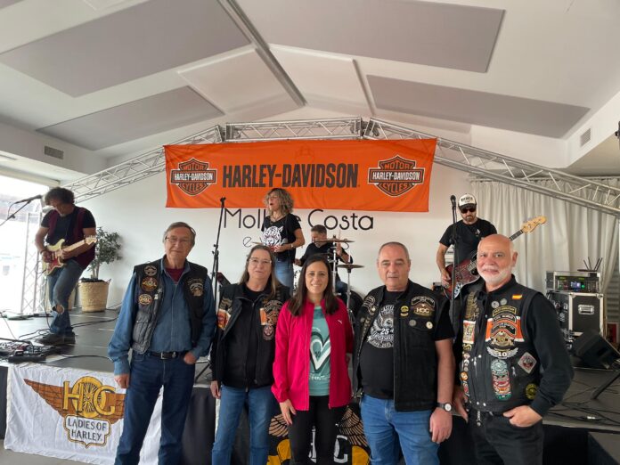 Group of Harley-Davidson riders posing on a stage with an orange Harley‑Davidson banner behind them.