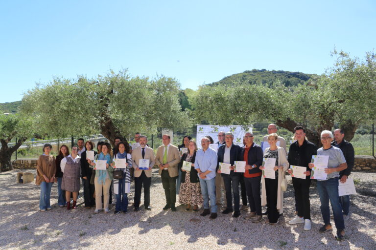 Group of people standing outdoors on a sunny day, many holding certificates or papers, posing for a photo in front of olive trees and hills.