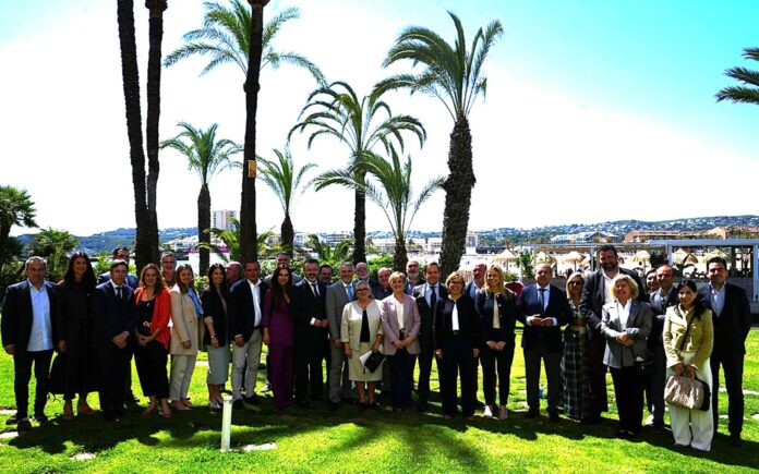 Group of businesspeople posing together on a sunny lawn with tall palm trees and a coastal city in the background.