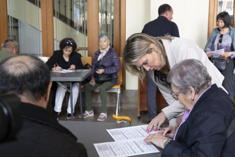 Seniors seated at a table filling out forms while a woman leans over to sign a document nearby.