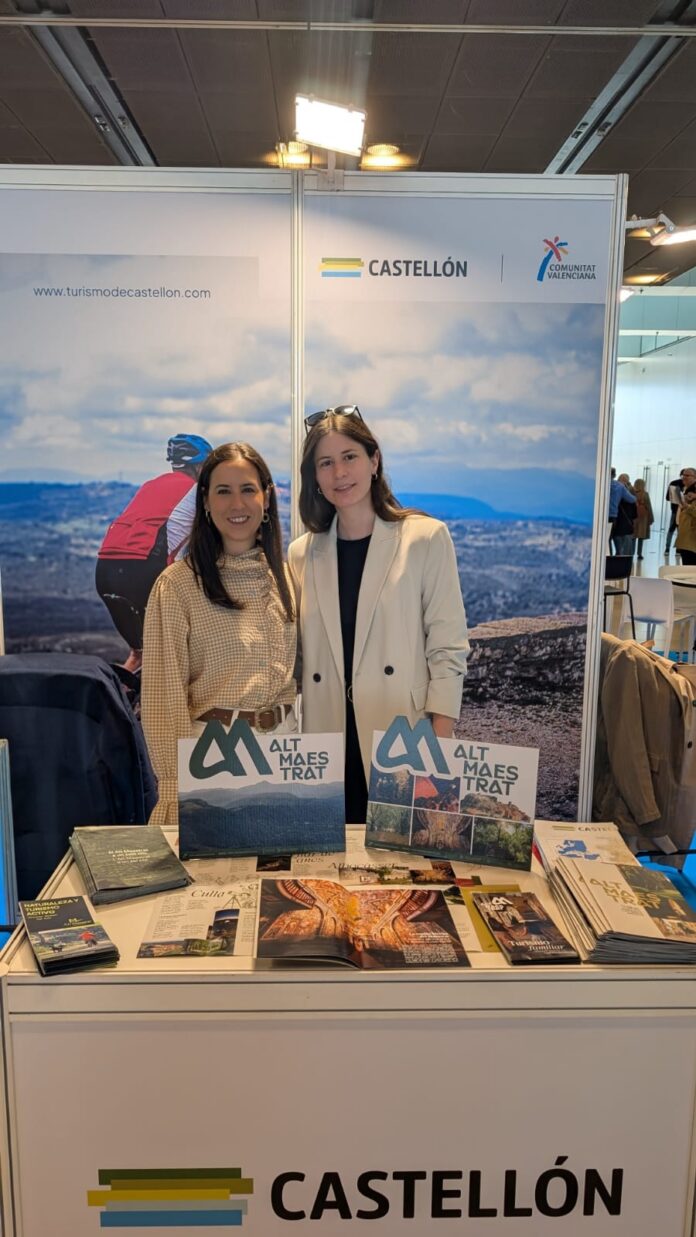 Two women stand at a Castellón tourism booth with brochures and a large backdrop showing a scenic landscape behind them.