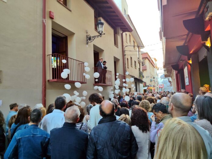 Crowded street festival with white balloons tied to balcony railings; spectators on balconies and below watch the event.