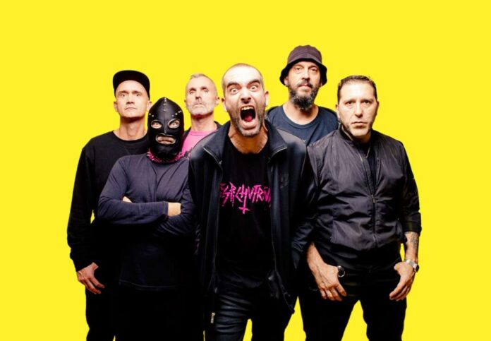 Six-member rock band posing together against a bright yellow backdrop, with one member wearing a mask in front row left.