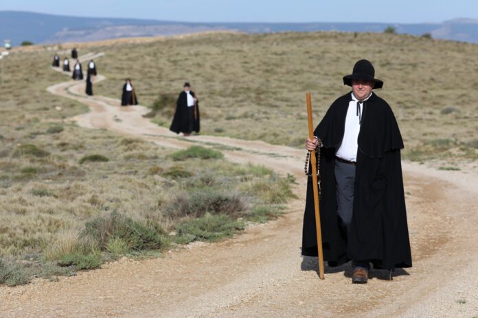 Man in a black cloak and wide-brimmed hat stands holding a wooden staff with a rosary in the desert, a dirt path winding behind him and others dressed similarly in the distance.