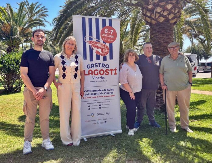 Five people pose outdoors beside a tall banner reading 'Gastro Llagostí' in Vinaròs, on a sunny day with palm trees in the background.