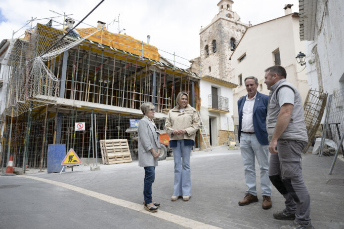 Four adults stand and converse on a street lined with a building under construction, scaffolding, and safety netting behind them.