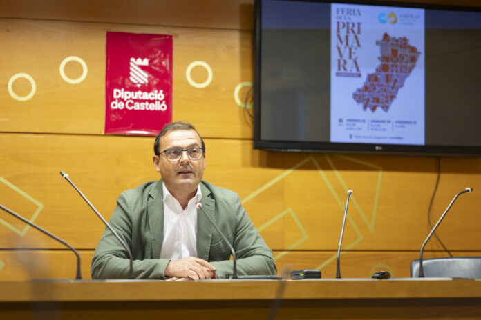 Man in a green blazer and glasses sits at a panel desk with microphones, wood-paneled wall behind and a red Diputació de Castelló banner to the left.