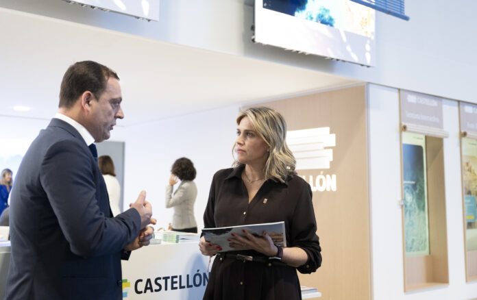 Two professionally dressed individuals talk at a marketing booth, woman holding brochures near a Castellón display.