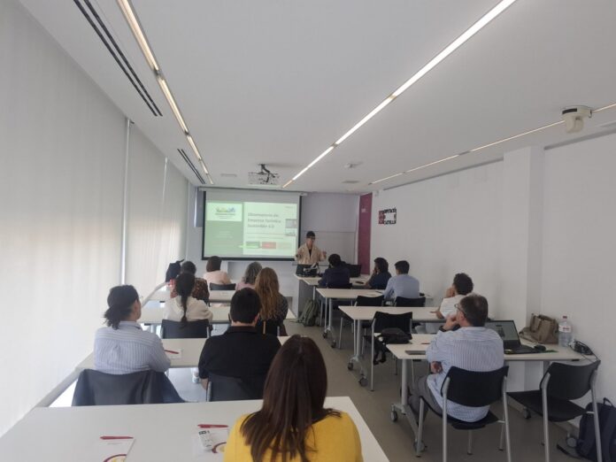 Speaker giving a presentation to a group in a bright classroom with a projected slide on screen at the front.