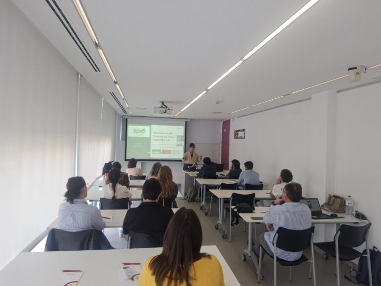 Speaker giving a presentation to a group in a bright classroom with a projected slide on screen at the front.