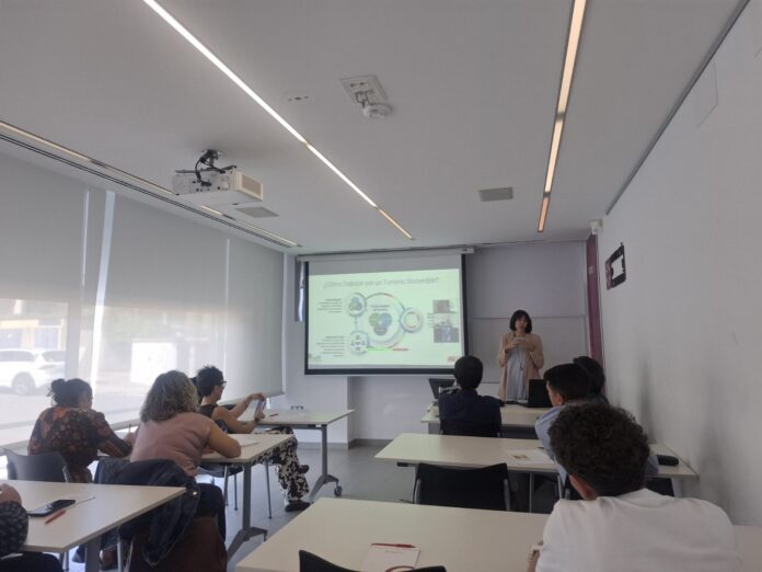 Teacher presenting a slide titled '¿Cómo Trabajar por un Turismo Sostenible?' to students in a bright classroom with a projector screen at the front.