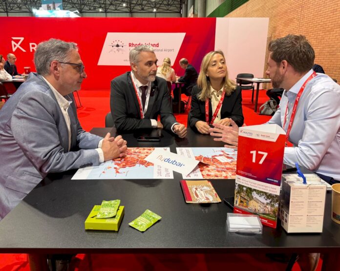 Four professionals in a conference booth discuss at a table with maps and brochures on display behind them in a red trade-show area.