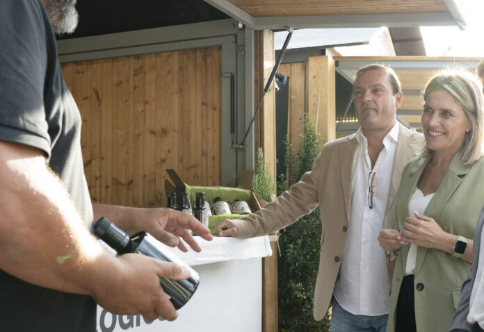 People at an outdoor wine stall exchanging a bottle, with a man in a beige blazer and a smiling woman nearby.
