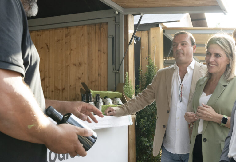 People at an outdoor wine stall exchanging a bottle, with a man in a beige blazer and a smiling woman nearby.