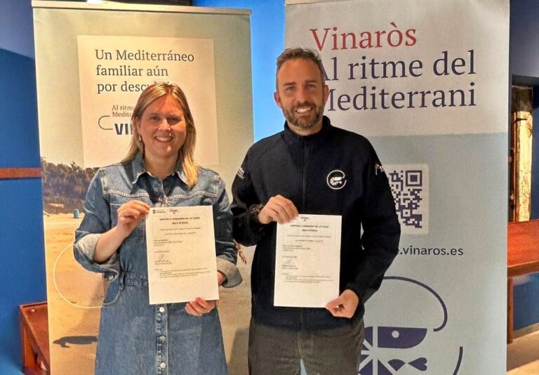 Two people stand smiling, each holding a document, in front of banners for Vinaròs Mediterranean rhythm festival.