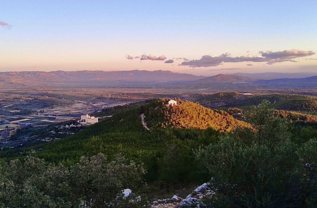 Sunset-lit valley with rolling hills, distant mountains, and a foreground of olive trees; a small white building sits among the trees on the lower left.