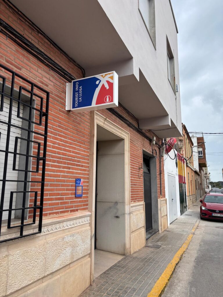 Blue and red 'Tourist Info La Losa' sign attached to a brick building along a narrow sidewalk street.