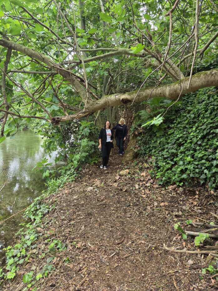 Two women walking along a dirt path beside a river under a large overhanging tree with green leaves at the top of the image.
