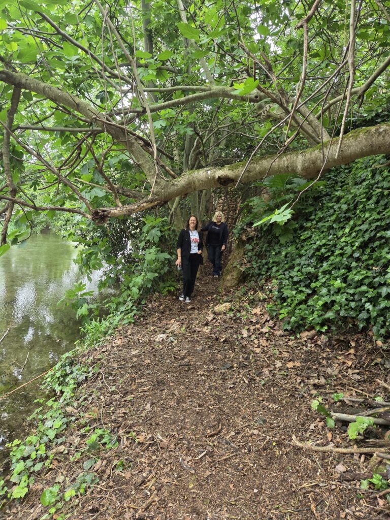 Two women walking along a dirt path beside a river under a large overhanging tree with green leaves at the top of the image.