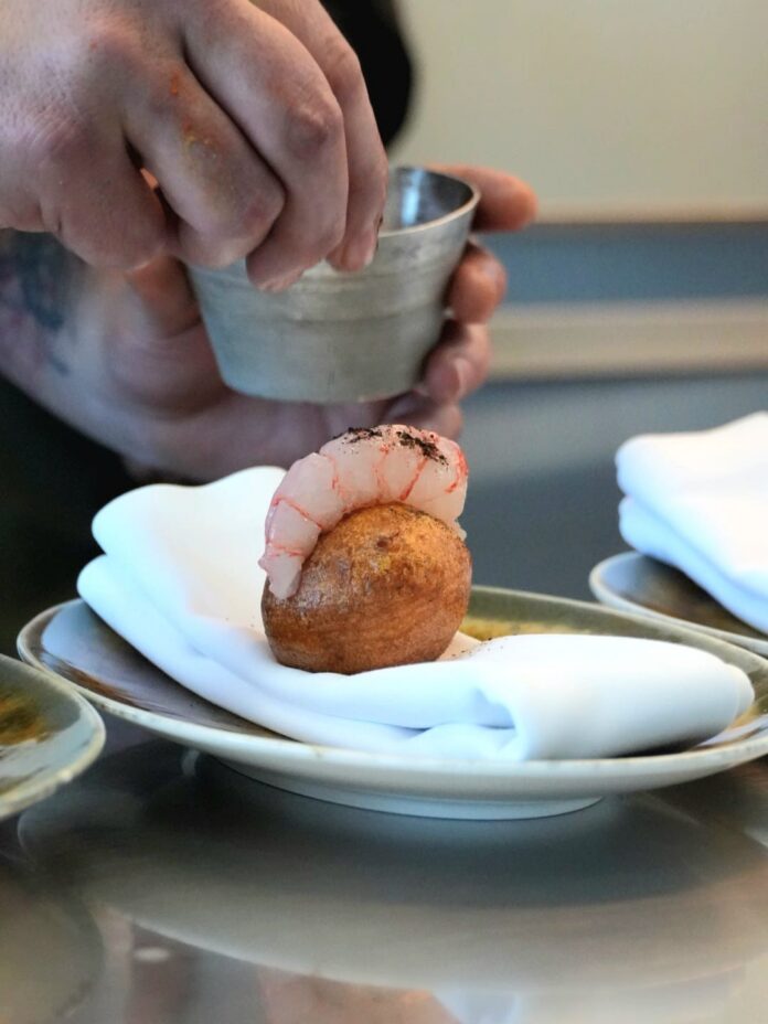 Shrimp curled on top of a small fried dough ball, with a server pouring sauce from a metal cup above it.