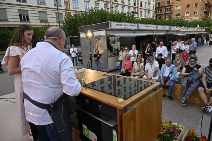 Chef wearing a white apron cooks at a large grill on a wooden counter as a crowd watches in an outdoor market area.
