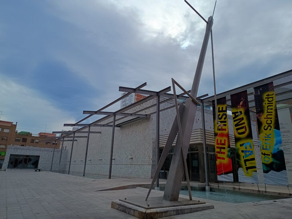 Outdoor plaza with intersecting gray metal beams sculpture next to colorful vertical banners by a white brick building.