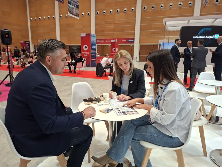 Three professionals sit at a white round table reviewing documents at a conference venue, expo booths visible in the background.