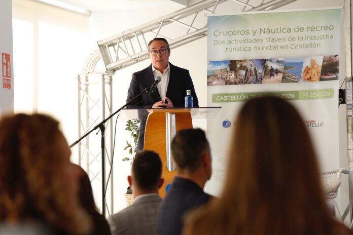 Man in a suit giving a talk at a wooden podium with a microphone, audience in foreground and a promotional banner behind him.