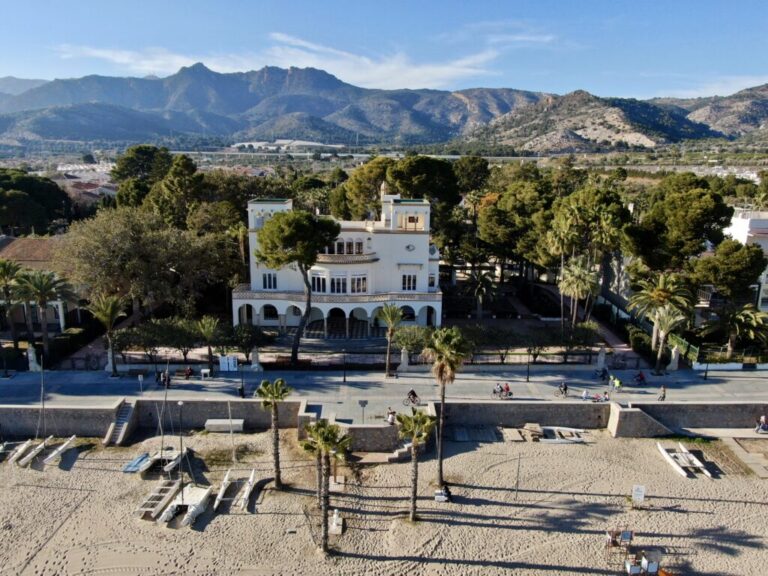A white villa along a palm‑lined promenade on a sandy beach, with boats and distant mountains beyond the shore.