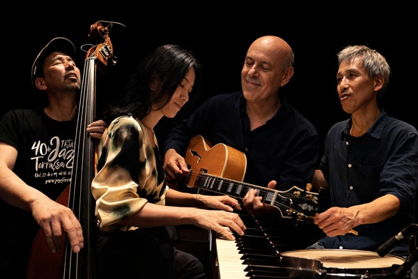 Four musicians perform together in a dark studio: bassist, pianist, guitarist, and drummer in a collaborative jazz jam.